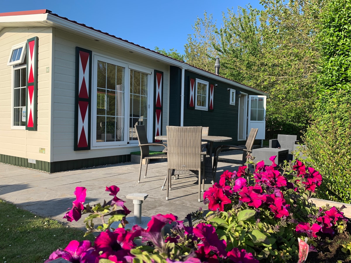 A charming chalet exterior is set against a clear blue sky, showcasing large windows and decorative shutters. A patio area features a dining table with four chairs, surrounded by vibrant pink flowers and greenery, creating a serene outdoor space.