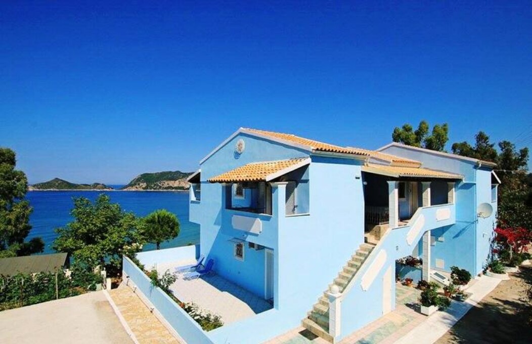 A blue house stands on a hillside, showcasing multiple levels with a staircase leading to the entrance. Lush greenery surrounds the property, while the deep blue sea is visible in the background against a clear sky.