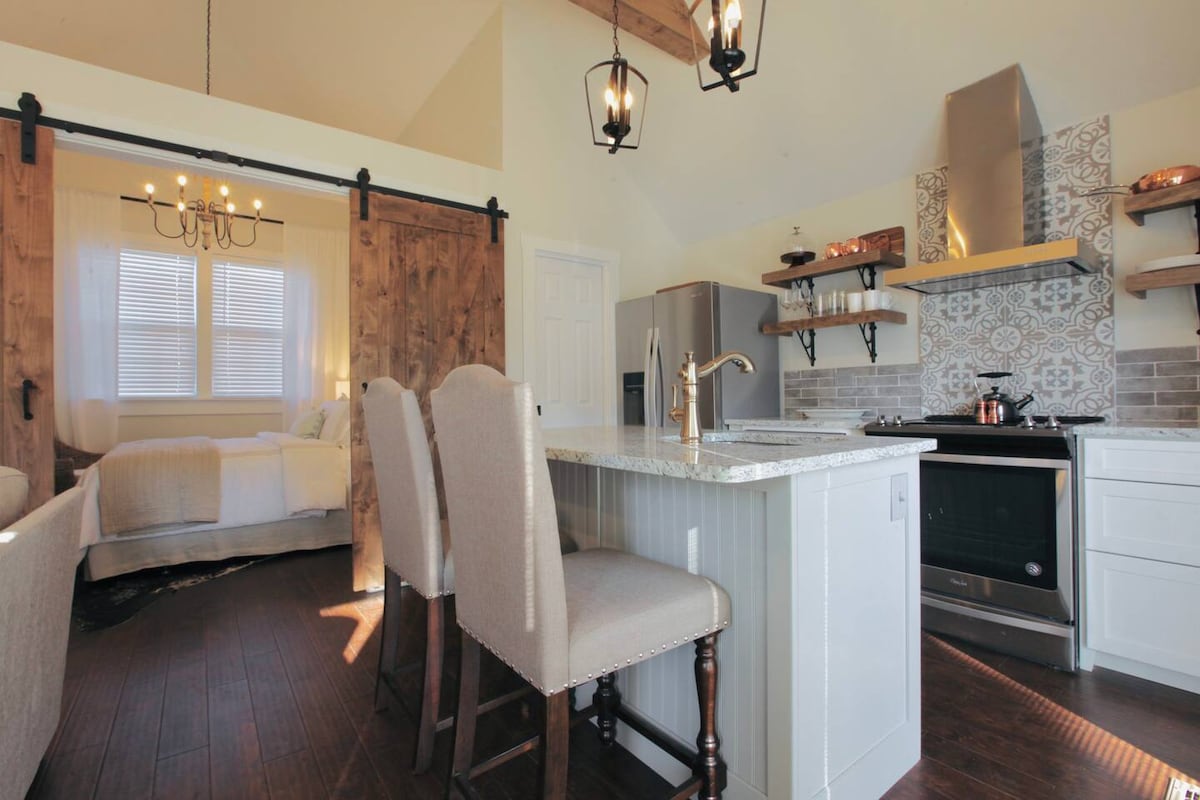 A modern kitchen area features a spacious island with seating for four. Rustic wood accents are complemented by light cabinetry and decorative tile backsplash. Natural light filters through large windows, illuminating the open layout that connects to a cozy sleeping area.