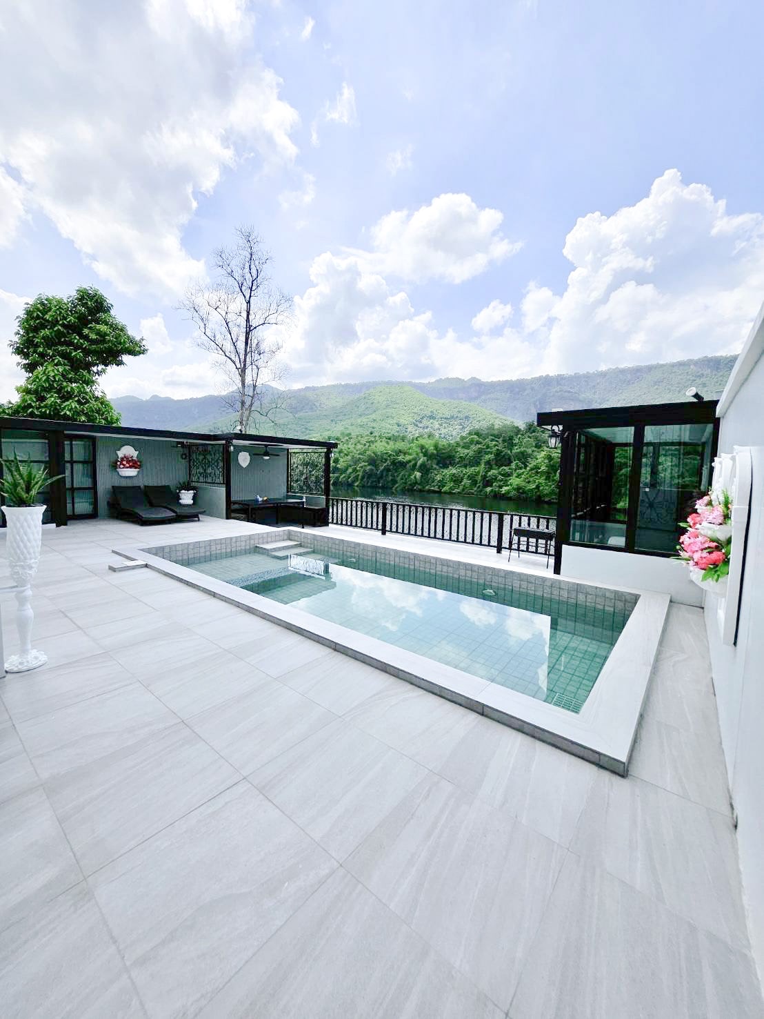 An outdoor area features a modern swimming pool surrounded by stone tiles. Lush greenery and mountain views are visible in the background. Comfortable seating is arranged nearby, with a glass-enclosed space offering a panoramic view of the scenery.