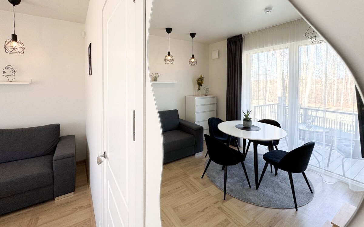 A modern living and dining area is featured, showcasing a round table surrounded by four black chairs. Two gray sofas are positioned against the walls, and natural light fills the space through large windows adorned with sheer curtains. Subtle decorative elements are present on the shelves.