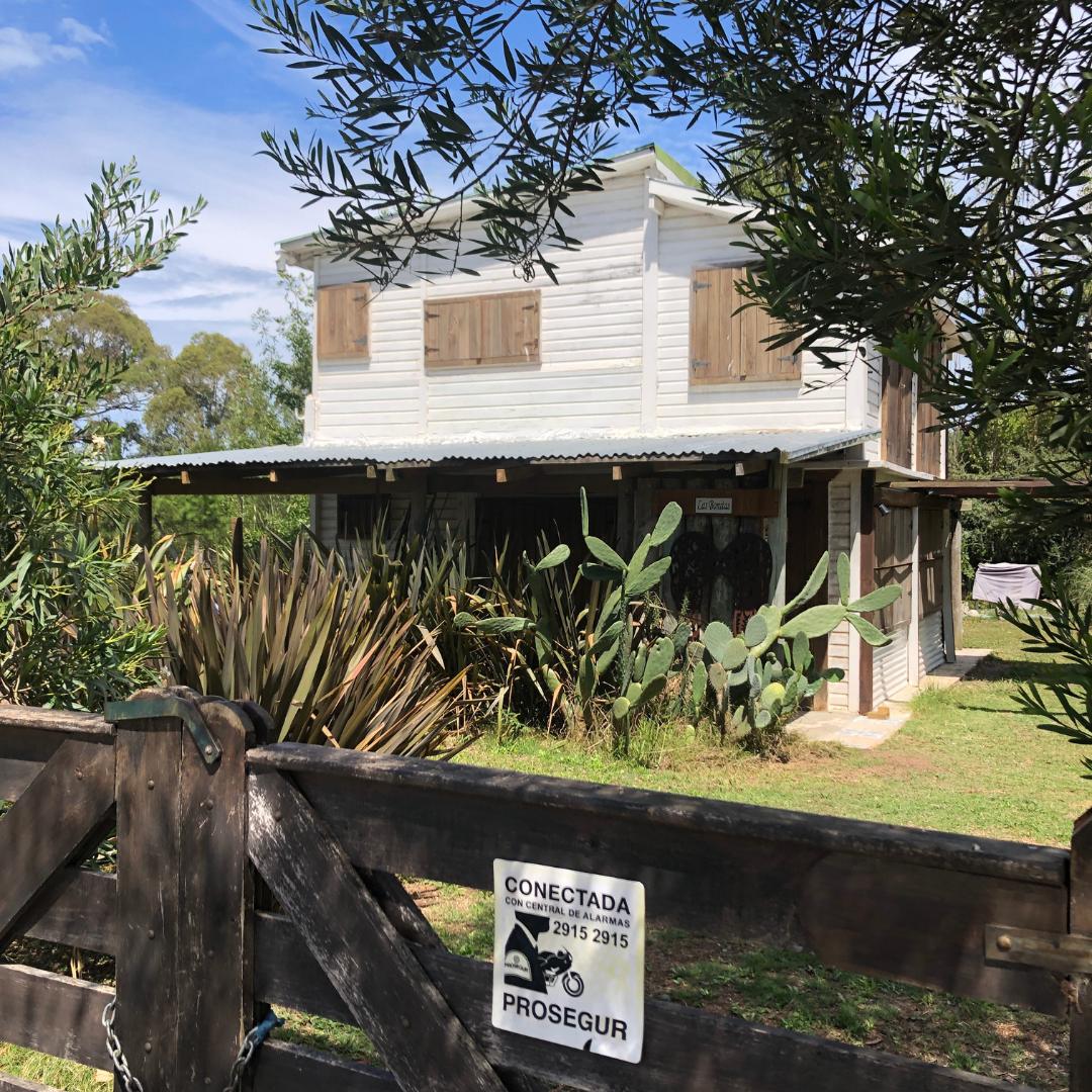 A two-story white house with wooden shutters is framed by lush greenery, including succulents and tall plants. The front gate is made of wood and features a security sign. Sunlight filters through the surrounding trees, highlighting the outdoor setting.