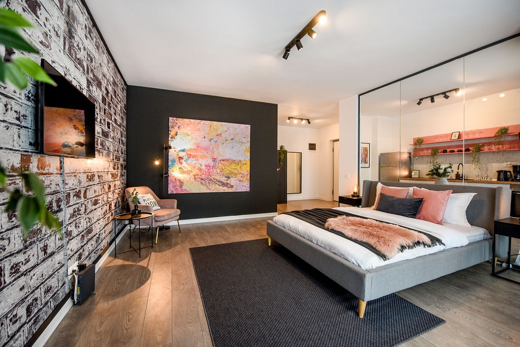 A spacious bedroom features a comfortable bed with a light-colored duvet, complemented by decorative pillows. A large abstract art piece adorns the wall, while a stylish armchair and small table are positioned nearby. Soft light is provided by wall-mounted fixtures, contributing to a serene ambiance.