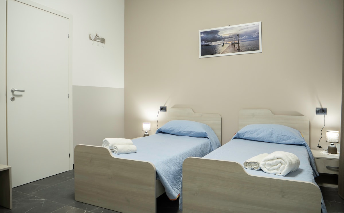 A neatly arranged sleeping area features two spacious beds with soft blue linens. Each bed is complemented by a bedside table with lamps. A simple photograph hangs above the beds, and a door is positioned to the left, leading out of the room.