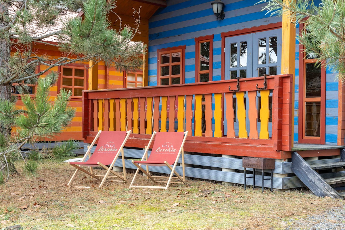 Two canvas chairs are positioned on a wooden deck, showcasing vibrant colors of orange and blue from the nearby cabin. Surrounding evergreen trees add a natural element to the outdoor space, creating a serene environment.