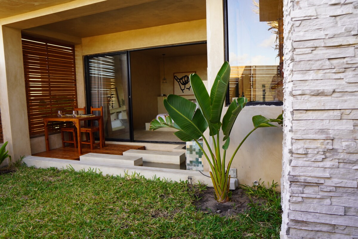 A modern beachfront unit features a large sliding glass door leading to a private outdoor area. Lush greenery surrounds the entrance, with a low step and a small dining area visible. Natural light fills the interior space, enhancing a sense of openness.