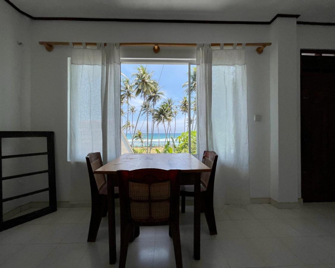 A dining area features a wooden table surrounded by four chairs, set against a backdrop of large windows with sheer white curtains. The ocean and palm trees are visible through the window, enhancing the bright and open atmosphere of the space.