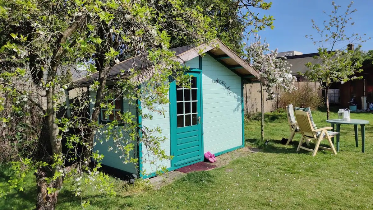 A quaint cabin with a turquoise exterior is set within a lush garden, framed by blooming trees. The entrance features a decorative door with glass panels. Outdoor seating is provided with garden chairs and a small table nearby, inviting guests to enjoy the serene surroundings.