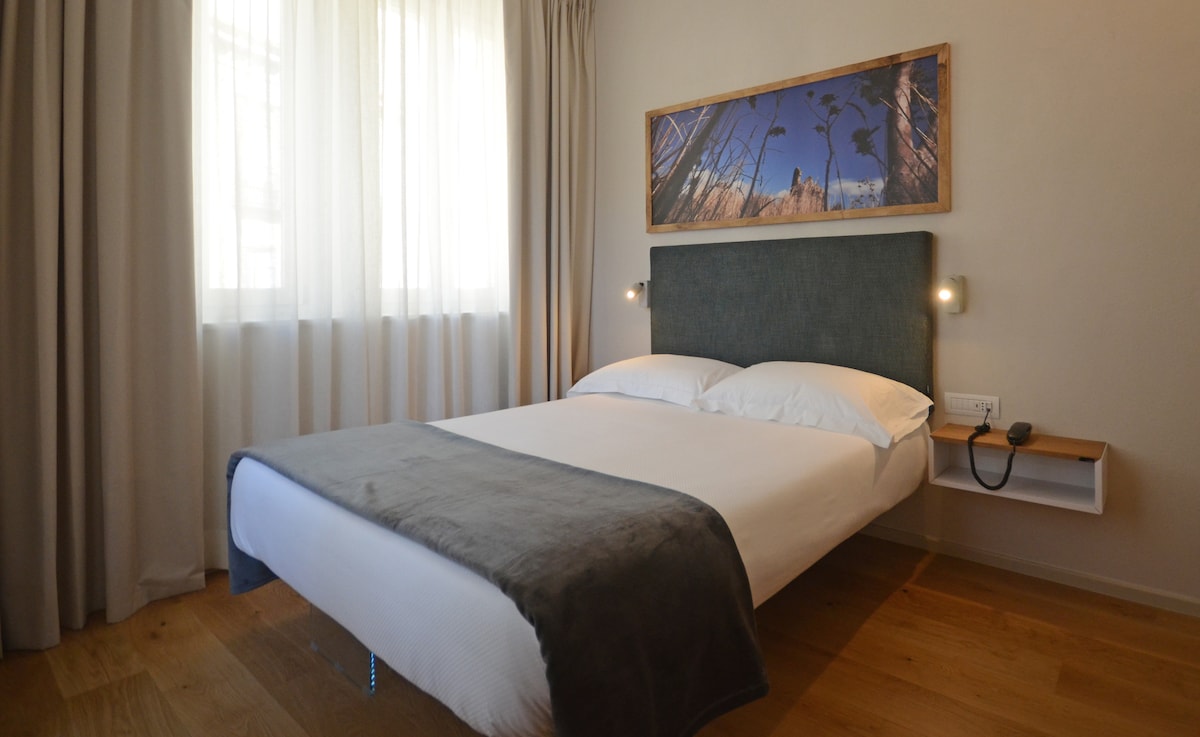 A spacious sleeping area is highlighted by soft natural light filtering through sheer curtains. The bed is framed by a textured headboard, complemented by a simple wall-mounted lamp on either side. A wooden nightstand with minimalistic decor is positioned beside the bed.