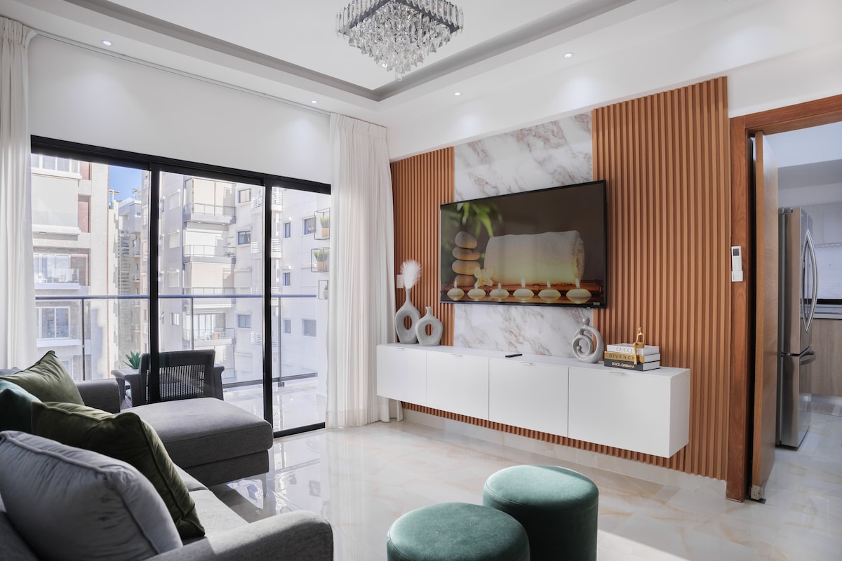 A modern living space is showcased with a spacious seating area featuring a gray sofa and two green stools. A wall-mounted flat-screen TV is located above a sleek white media console. Natural light is visible through large sliding glass doors leading to a balcony.