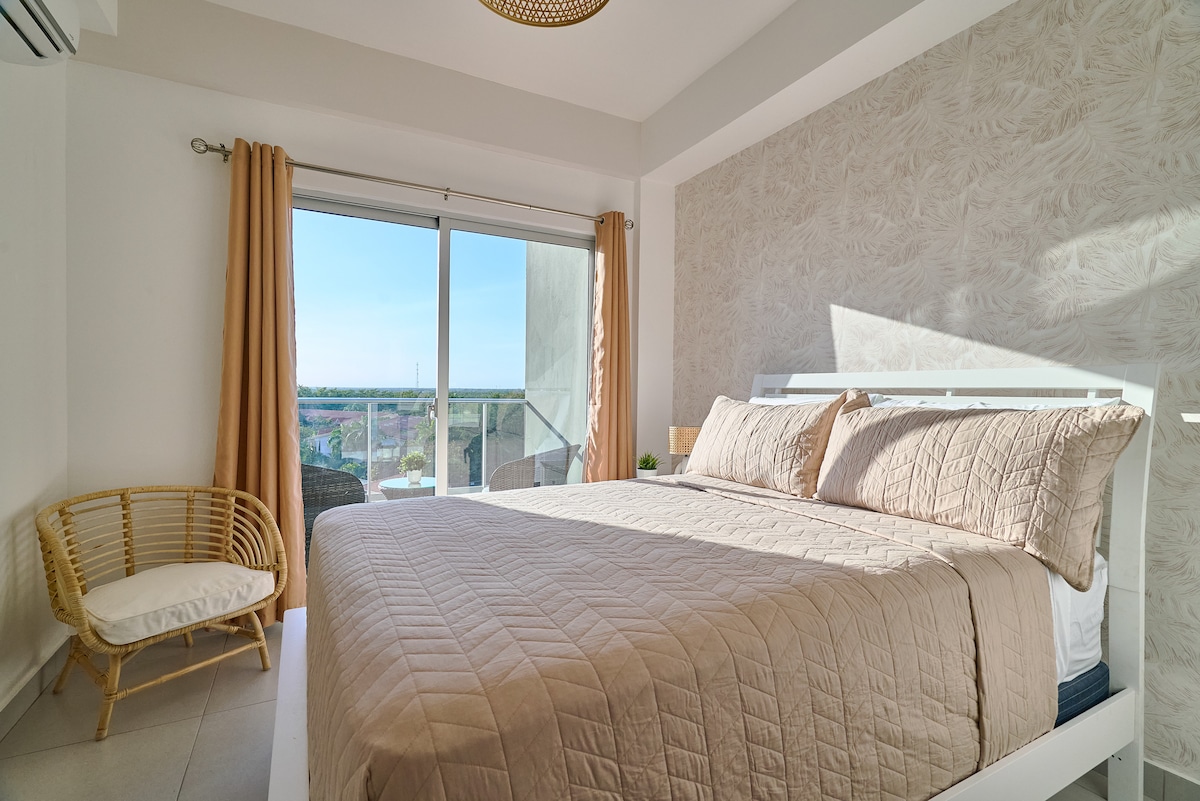 A spacious bedroom features a large bed dressed in soft beige linens. Natural light floods the room through wide sliding glass doors, which open to a balcony. A comfortable chair rests nearby, complementing the clean and modern decor.