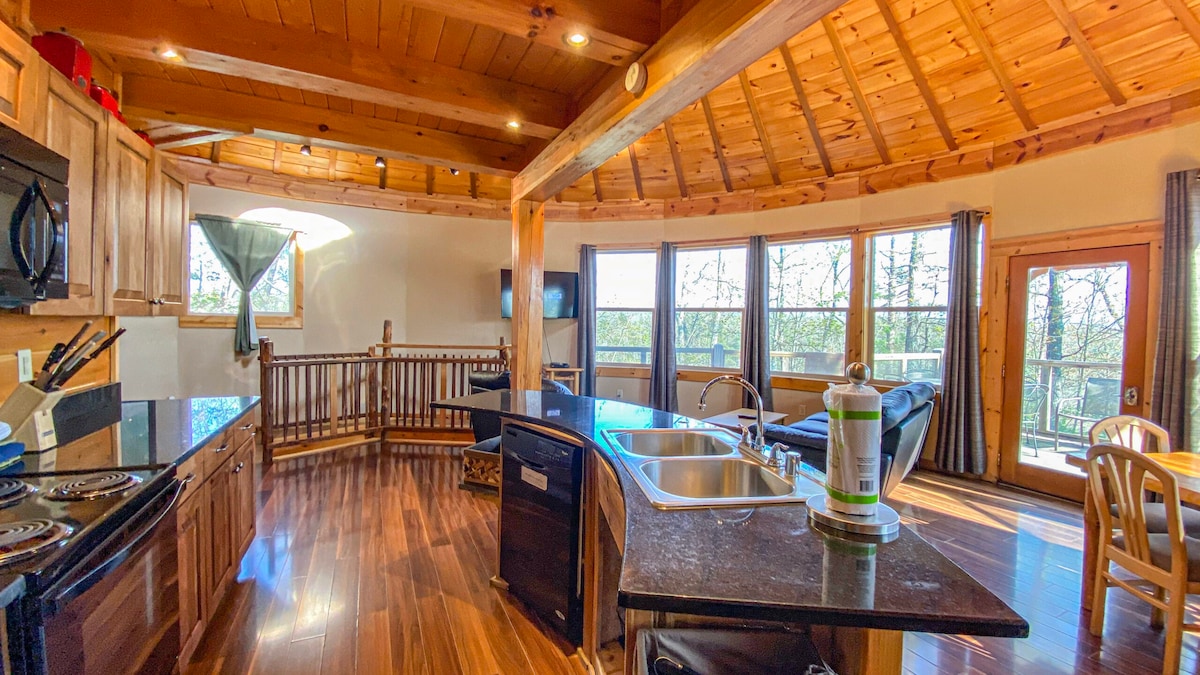 The spacious kitchen and living area features a wooden ceiling with exposed beams. Large windows flood the space with natural light and offer views of the surrounding wooded landscape. A central island with a kitchen sink is surrounded by wooden cabinetry, while a comfortable seating area is visible nearby.