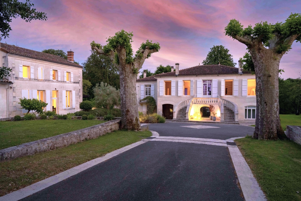 The exterior of the Maison du Gardien is showcased, featuring elegant architecture under a vibrant sky at dusk. The driveway leads to the front entrance, framed by two trees. Light spills from the windows, highlighting the inviting atmosphere of the property.