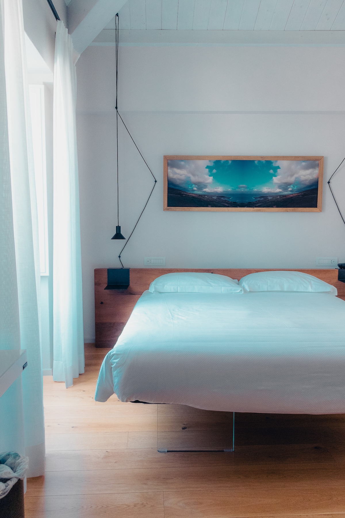 A bright sleeping area features a spacious bed with a white duvet and two pillows. Natural light enters through a large window draped with light curtains. Beside the bed, pendant lamps provide additional lighting, while a landscape artwork hangs above the headboard.