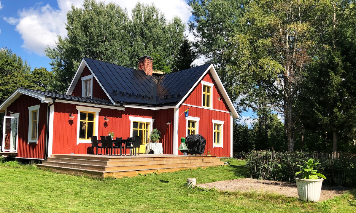 A charming red cottage features a dark roof and yellow-framed windows, surrounded by greenery. A spacious deck is visible, furnished with a black grill and a dining set, perfect for outdoor meals. Lush trees provide shade along the property.