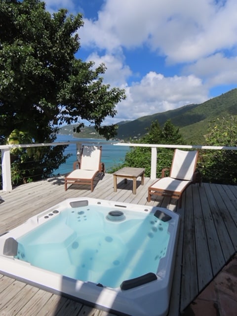 An outdoor deck offers a hot tub surrounded by lush greenery and partial views of the ocean. Two lounge chairs are positioned nearby, accompanied by a small table. The scene is illuminated by bright blue skies and scattered clouds.