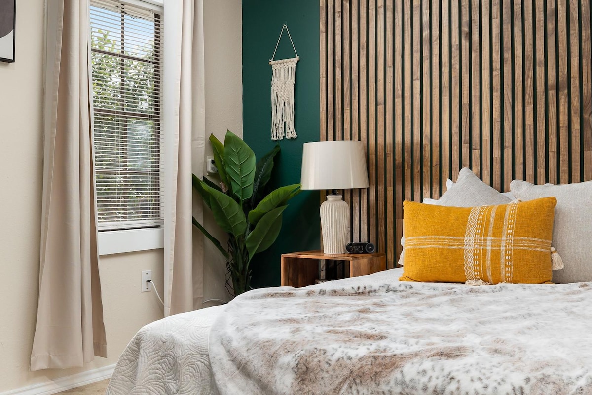A cozy bedroom featuring a spacious bed covered with a soft, textured blanket. A decorative yellow pillow adds a pop of color. In the background, a wooden accent wall and a greenery plant provide a calming effect, while natural light filters through a window dressed with light curtains.