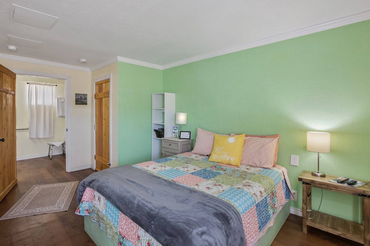 A cozy bedroom is presented with a spacious bed featuring a colorful quilt. Soft green walls enhance the tranquil setting, while a bedside lamp and a small nightstand add practicality. Natural light fills the room from a partially open curtain, creating a warm environment.
