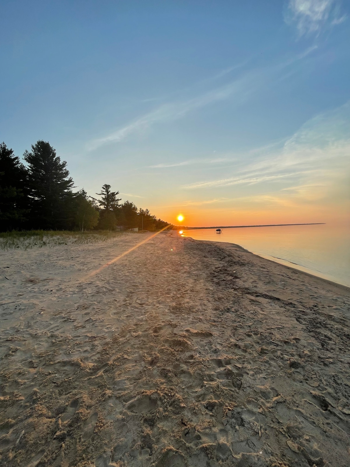 A sandy beach extends towards a calm lake, where the sun is setting on the horizon. Tall trees line the shore, with the sky displaying soft hues of orange and blue. The water reflects the colors of the sunset, creating a serene and peaceful scene.