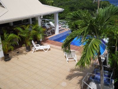 An outdoor terrace is shown, featuring a private swimming pool surrounded by lounge chairs and potted palm trees. The covered area includes dining furniture, creating a space for relaxation and gatherings. Views of the lush landscape and distant water are visible in the background.