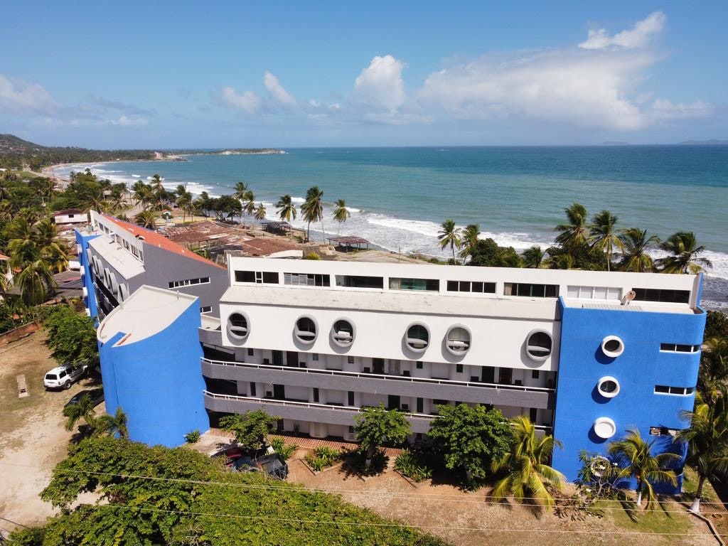 A modern building is viewed from above, featuring a mix of blue and grey exterior. Palm trees are situated around the property, and the ocean is visible in the background, showcasing gentle waves. The surrounding area indicates proximity to the beach.