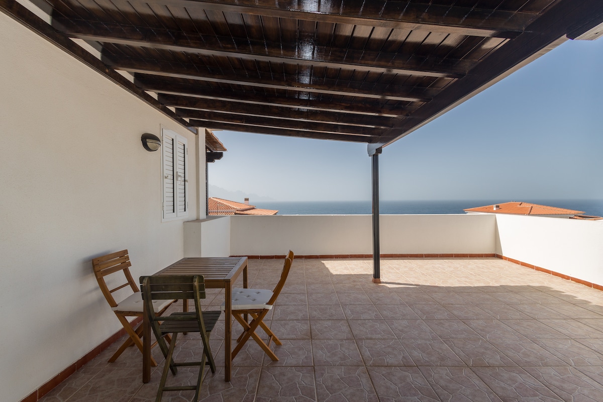 A spacious terrace is featured with a wooden overhang, providing partial shade. Two folding chairs and a small table are positioned, offering a place to relax and enjoy the coastal views beyond. The ocean can be seen in the distance, creating a serene backdrop.