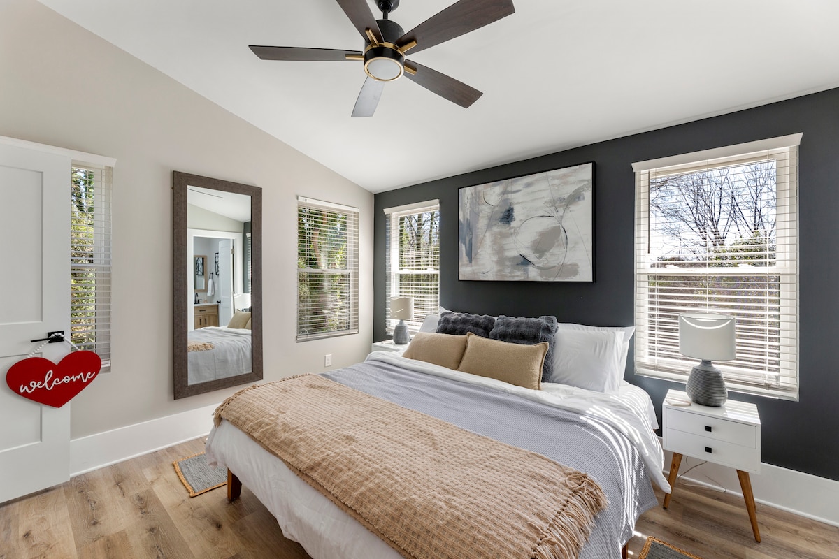 The spacious bedroom is complemented by large windows allowing ample natural light. A comfortable bed with layered linens occupies the center, while a full-length mirror and a nightstand add functional elements to the layout. A ceiling fan provides airflow, enhancing the room's comfort.