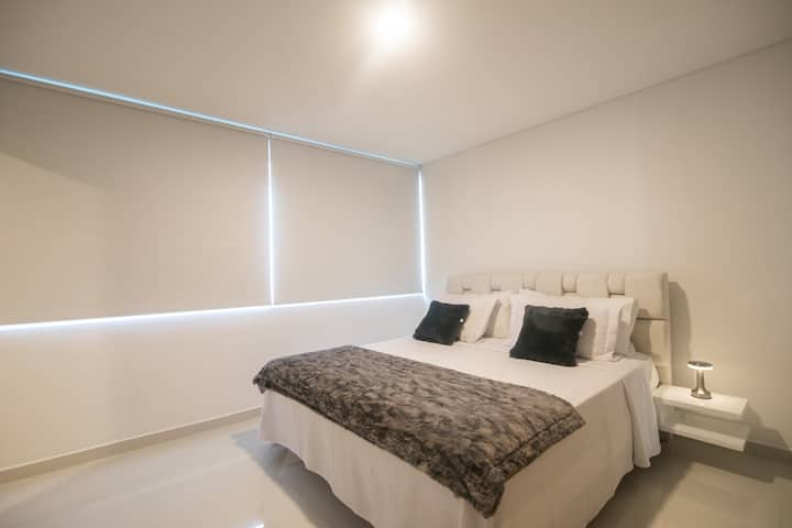 Spavaća soba 2 na gornjoj etaži s bračnim krevetom i sjenilima za zamračivanje; kupaonica nije vidljiva na slici. Pogled kroz prozor na plažu i obalu. 