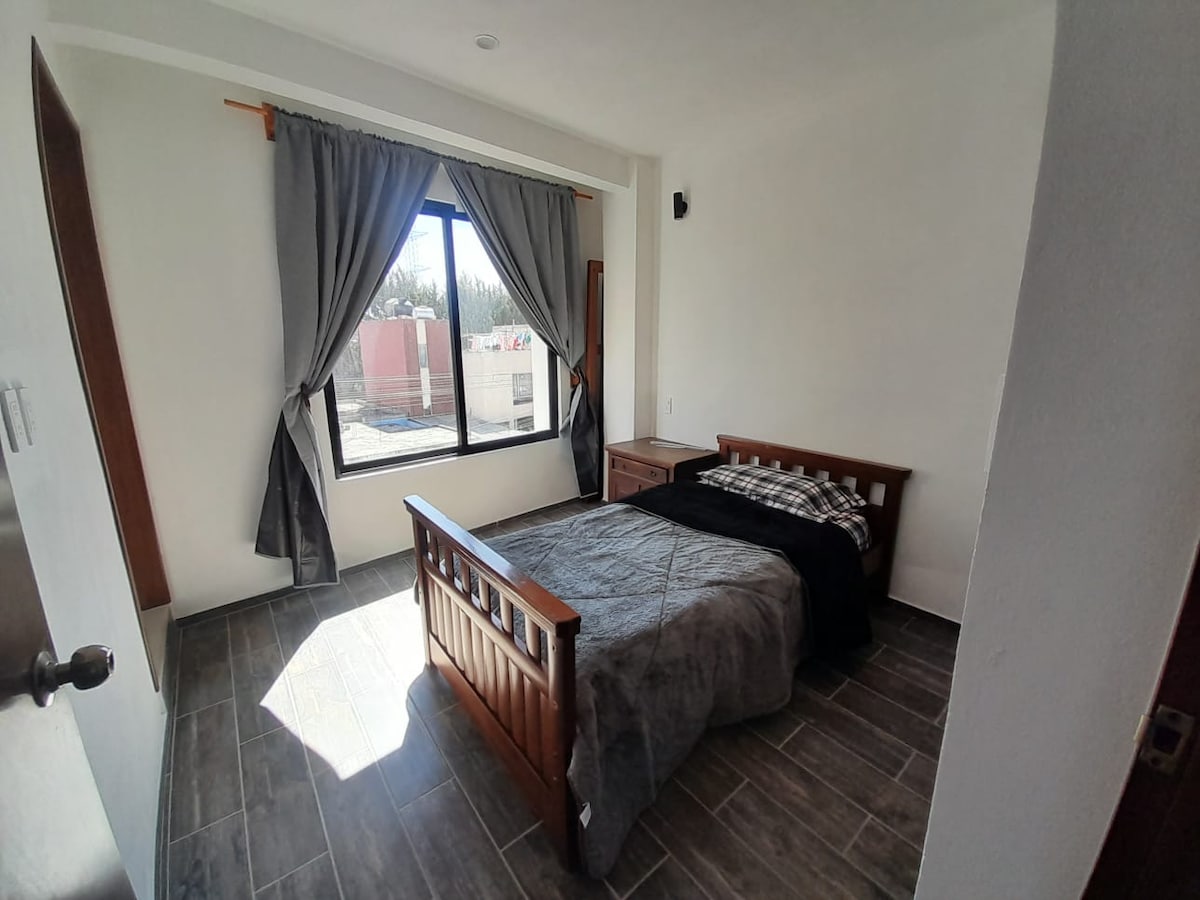 A well-lit bedroom features a wooden bed adorned with a dark quilt. Large windows allow natural light to fill the space, with gray curtains framing the view. Wooden accents are present in the furniture, contributing to a clean and organized ambiance.