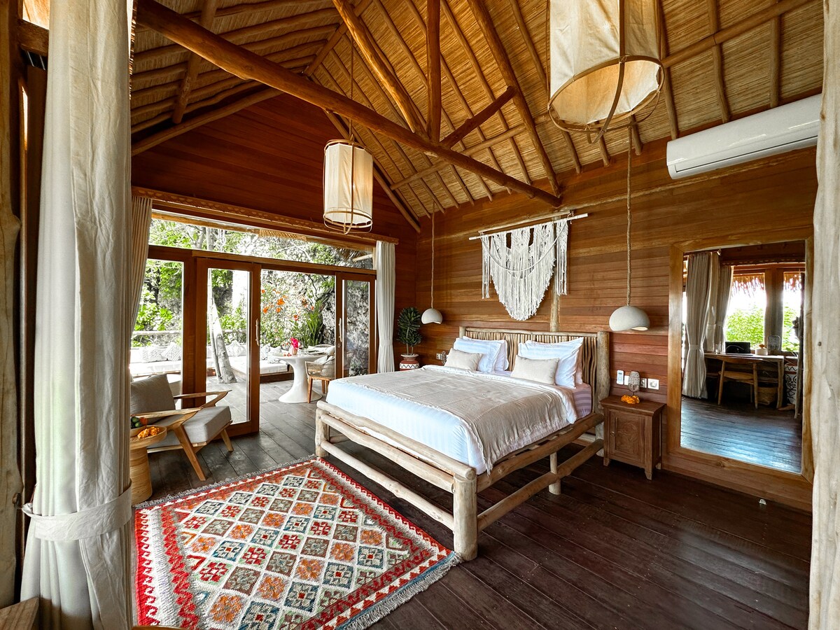 A cozy villa interior features a spacious king-sized bed framed by natural wooden walls and a high thatched roof. Large glass doors lead to a private outdoor area, while a patterned rug adds warmth. A mirror reflects the inviting ambiance of the room.