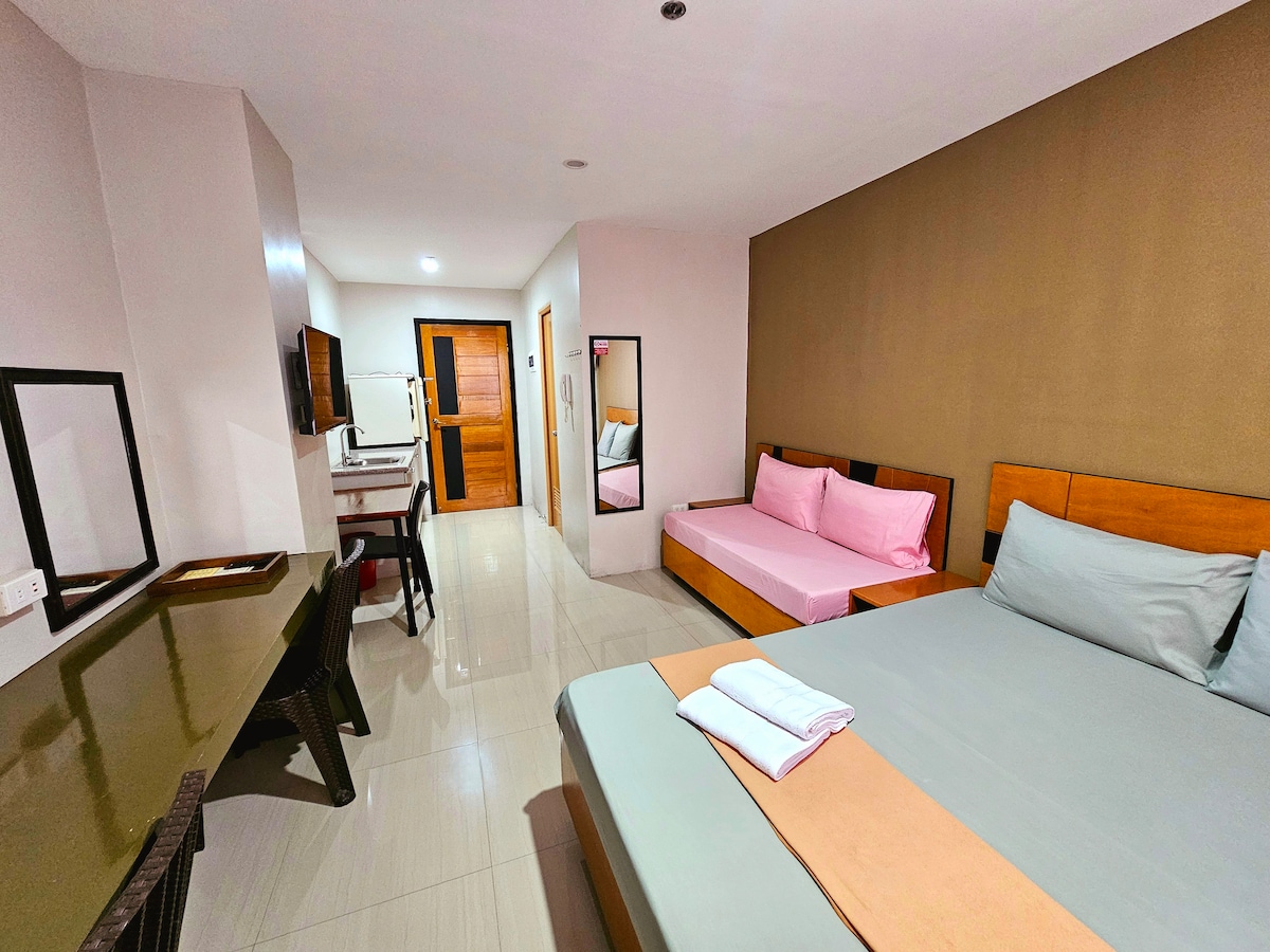 A spacious studio layout features a queen-sized bed with a neutral bedding arrangement alongside a single sofa bed. A large work desk sits adjacent to a wall-mounted television. The room is illuminated by natural light from the window, creating a welcoming environment.