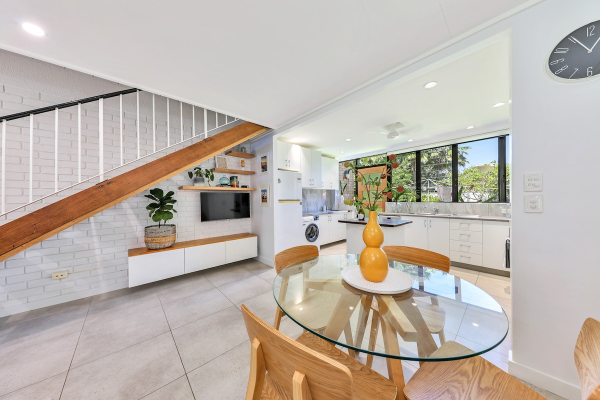 The open living and dining area features a round glass table with wooden chairs, illuminated by natural light from large windows. The kitchen is equipped with modern appliances and white cabinetry. A decorative plant provides a touch of greenery, enhancing the inviting atmosphere.