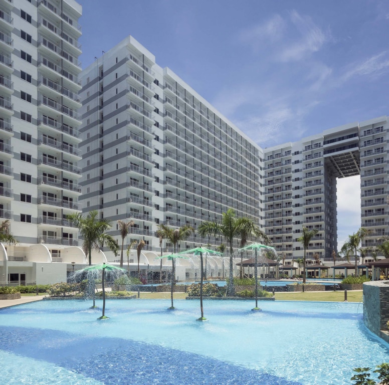 A serene outdoor swimming pool is surrounded by modern condominium buildings. Clear water reflects sunlight, and decorative fountains are positioned within the pool. Lush greenery and palm trees enhance the ambiance, providing a refreshing atmosphere for relaxation.