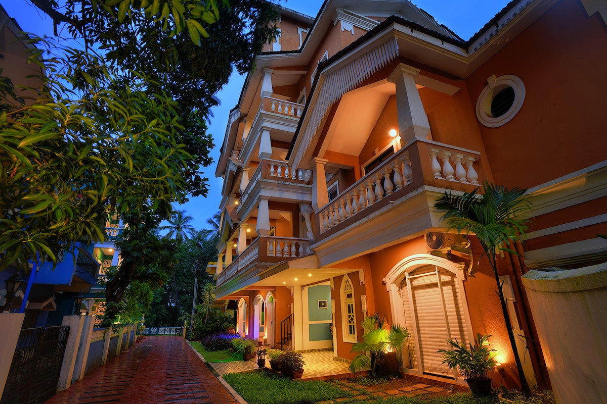 The exterior of the building showcases Indo-Portuguese architecture, with multiple levels featuring balconies and elegant railings. Lush greenery surrounds the pathway, which is lined with subtle lighting, creating a welcoming atmosphere. The warm hues of the facade are complemented by the evening sky.