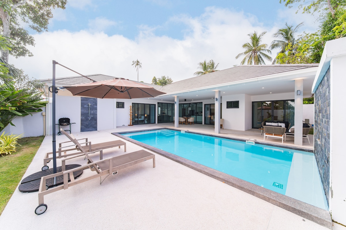 A modern villa exterior showcases a private pool surrounded by lounge chairs. An umbrella provides shade near the poolside. Large glass doors offer views into the spacious living area, while lush greenery enhances the tranquil setting.