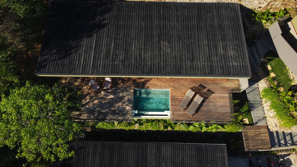 An aerial view illustrates a modern villa with a sleek black roof. An infinity pool is positioned adjacent to a spacious wooden deck, furnished with lounge chairs. Surrounding greenery enhances the serene environment, while the overall layout emphasizes privacy.