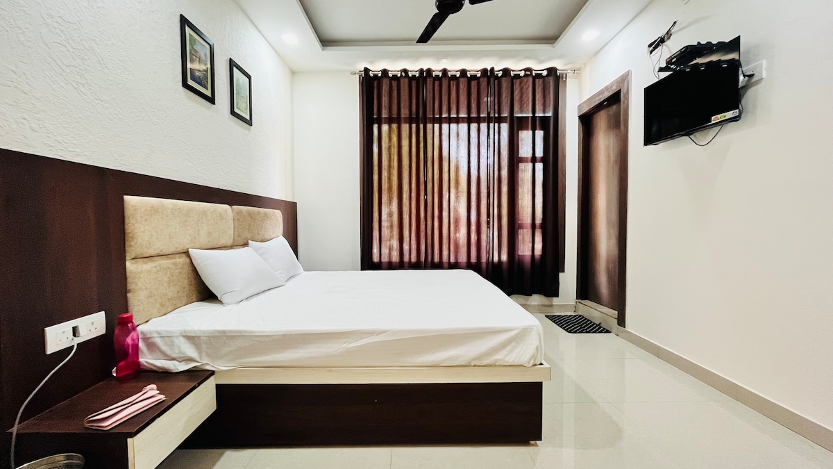 A spacious bedroom features a soft, neutral-toned bed with crisp white linens. Large windows with dark curtains allow natural light to filter through, illuminating the light-colored walls. A television is mounted on the wall, and a small table is located beside the bed.
