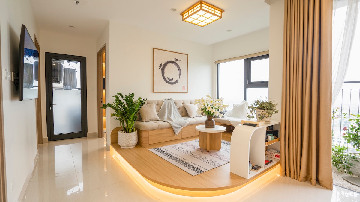 A bright living area is featured with a large, minimalist sofa adorned with cushions, facing a round coffee table. Elegant plants are placed on either side. A window allows natural light to fill the space, with sheer curtains softly draping to one side.