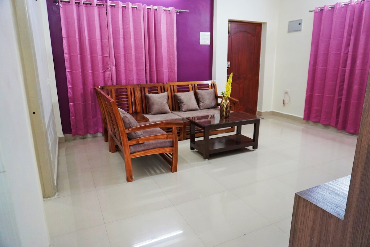 A living area features a wooden seating arrangement with plush cushions, positioned around a dark coffee table. Light-colored tiles reflect natural light, while purple curtains frame the windows, adding a pop of color to the neutral walls.