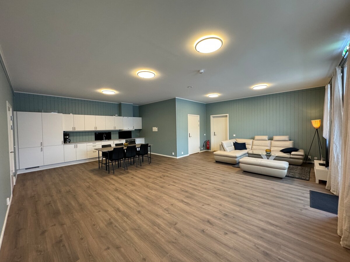 A spacious living area features light wood flooring and pale blue walls. An arrangement of modern sofas is complemented by a floor lamp. A dining table with several chairs occupies one corner, while a kitchen area is visible with cabinetry and appliances.