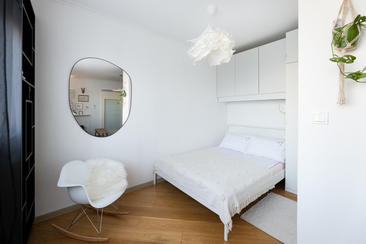 A minimalist sleeping area features a spacious bed with a light, textured coverlet. A unique, reflective wall mirror is mounted above a sleek chair, adding to the contemporary decor. Soft natural light fills the room, enhancing the bright white walls and warm wooden flooring.