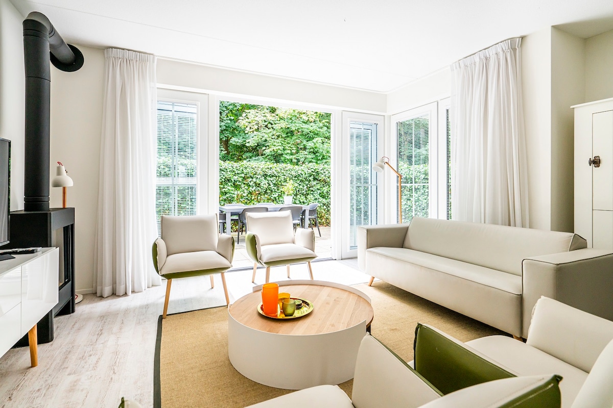 The living area features a light color palette with a spacious sofa and two accent chairs. Large glass doors open to a private patio, while natural light brightens the space. A round coffee table holds decorative items, enhancing the room's inviting design.