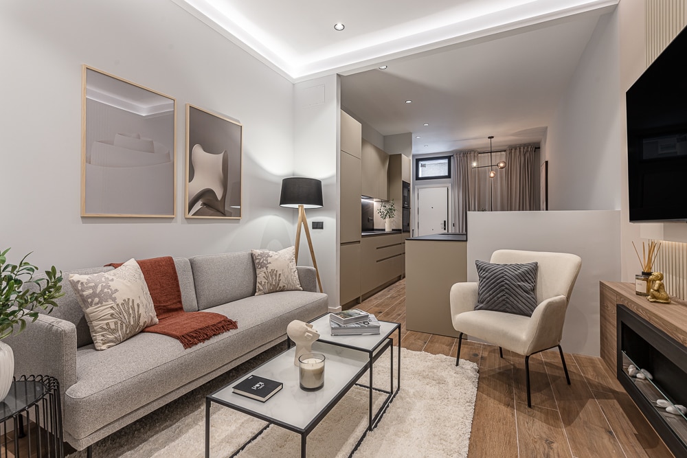 The living area features a light gray sofa adorned with accent pillows, accompanied by a modern armchair. A minimalist coffee table sits on a plush area rug, while framed art decorates the walls. Natural light fills the space, highlighting the warm wood flooring.