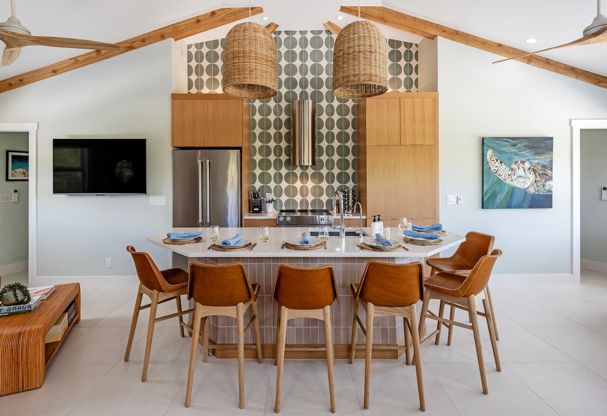 A spacious kitchen area features a large island with seating for six. The room showcases modern appliances, a stylish backsplash with circular patterns, and an open ceiling enhanced by natural wood beams. Light colors create a bright and inviting environment.