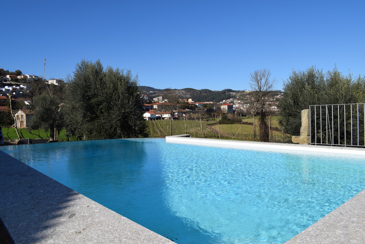 A serene outdoor infinity pool is depicted, featuring clear blue water that reflects the sky. Surrounding the pool, lush greenery and picturesque rural landscapes are visible, enhancing the tranquil setting. A stone border frames the pool, contributing to the peaceful ambiance.