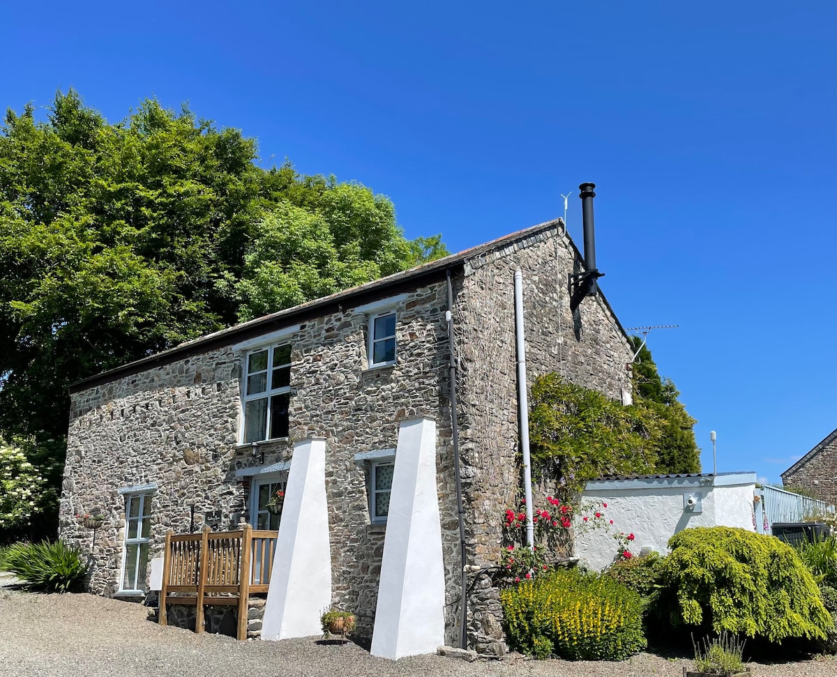 The exterior of Godrevy Barn is shown, featuring rustic stone walls and large windows that invite natural light. Lush greenery surrounds the structure, with a wooden deck area in front. Bright flowers add a splash of color to the peaceful rural setting.