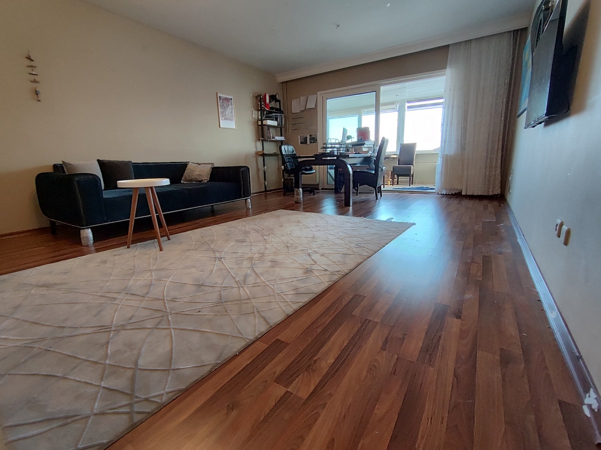 A spacious living area features warm wooden flooring and a soft, neutral rug at the center. A dark couch and small table are contrasted by a glass sliding door leading to a balcony. A workspace with a desk and chair is visible in the background.