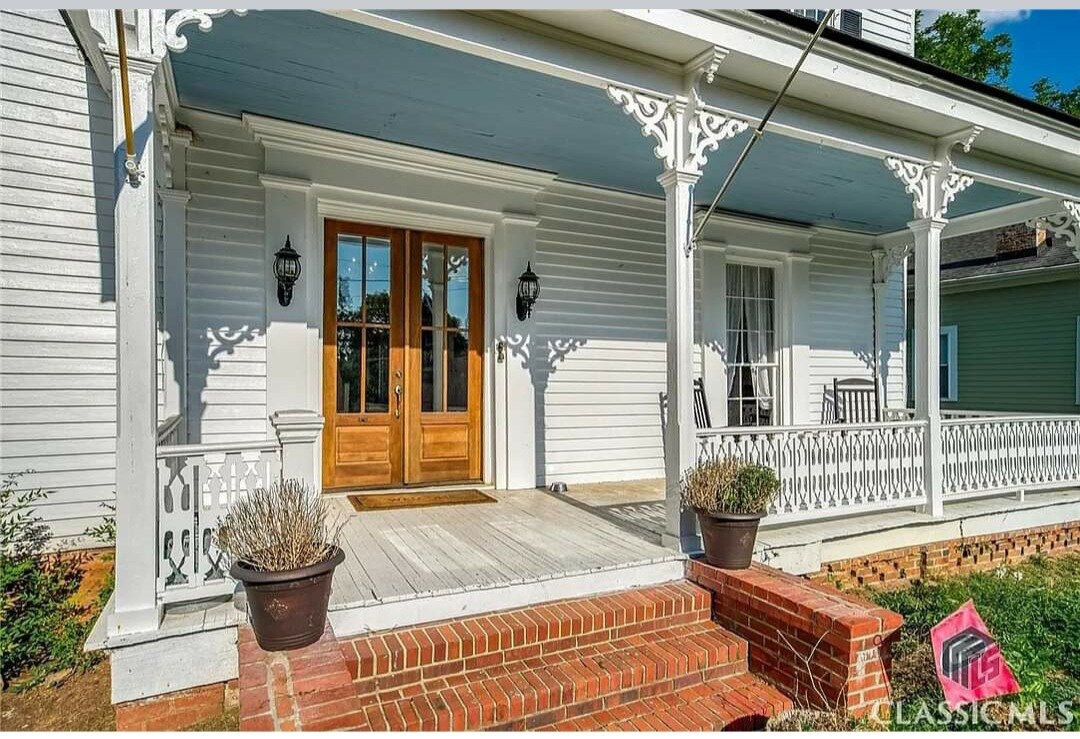 The front porch showcases classic colonial architecture, featuring a spacious wooden deck and elegant double doors. Decorative white railings provide a charming touch, complemented by planters. A brick walkway leads to the entrance, enhancing the welcoming appearance of the historic farmhouse.