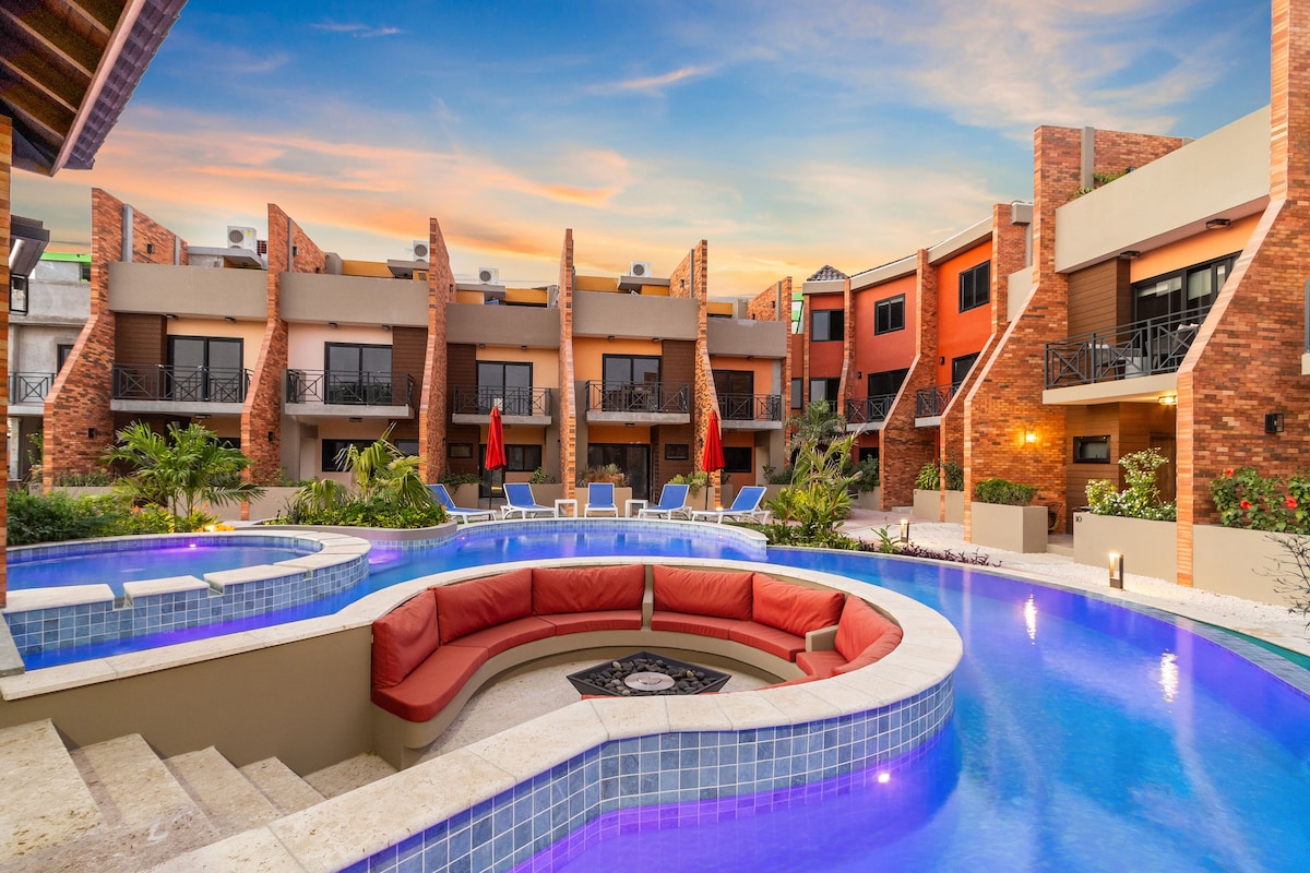 A resort-style pool area is showcased, featuring a circular lounge with bright red seating surrounding the water. Lush landscaping complements the surroundings, while townhouses provide a warm backdrop. Soft lighting reflects off the tranquil pool, enhancing the inviting ambiance of the space.