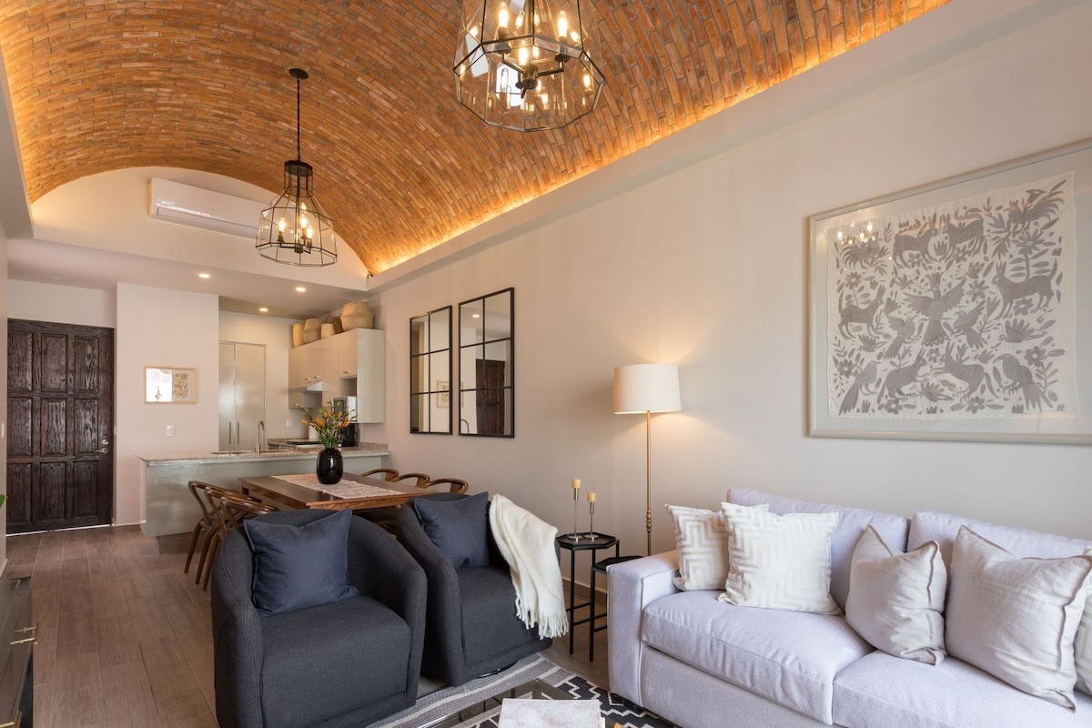 A spacious open-concept living area features a decorative vaulted ceiling with warm lighting. A soft sofa is complemented by two dark lounge chairs. A large dining table with elegant seating is visible in the background, along with kitchen amenities seamlessly integrated into the space.